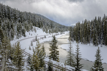 Empty Railroad Track Running alongside a River Winding its Way through a Snowy Forest on a Cloudy Winter Day. Banff National Park, AB, Canada.