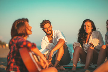 Happy friends sitting on the beach singing and playing guitar during the sunset