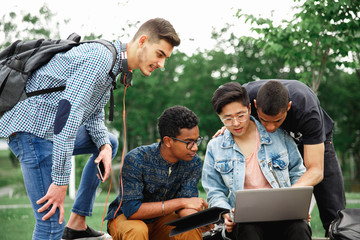 Group of young smiling students sitting on bench and working on laptop together while spending time in courtyard of university. Company of students studying outdoors