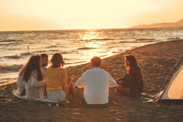 Happy friends sitting on the beach singing and playing guitar during the sunset