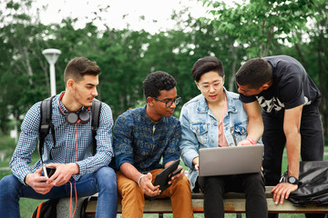 Group of young smiling students sitting on bench and working on laptop together while spending time in courtyard of university. Company of students studying outdoors
