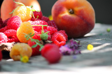 Still life with red and yellow raspberries and peaches. Fruit on a wooden table. Against the background of a bouquet of wildflowers. Close-up. Selective focus, side view, copy space.