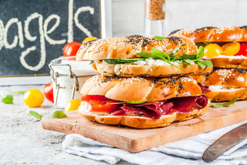Variety of homemade bagels sandwiches with sesame and poppy seeds, cream cheese,  ham, radish, arugula, cherry tomatoes, cucumbers, white grey textured background copy space