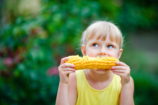 The beautiful girl bites corn on a farm.