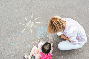 Top view of happy little girl and mother drawing with chalks on sidewalk. Caucasian female play together with kid preschooler with backpack outdoor. Mother and child activity. Good relationship