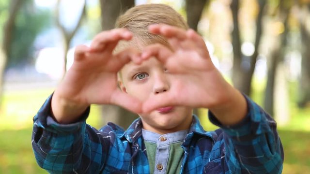 Young handsome blond boy forming heart shape with hands, looks through it into camera and smiles happily. Blurry city park in background. 