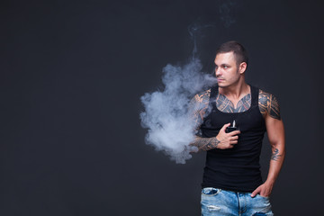 Vaper. The man with a muscular torso with tattoos smoke an electronic cigarette on the dark background