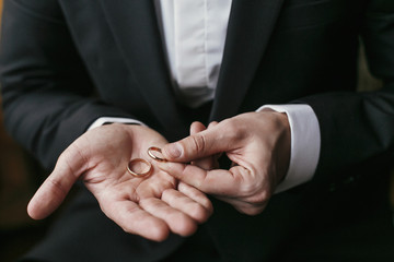 Wedding rings on palm hand. Groom in stylish suit holding golden wedding rings in hands, sitting in the room. marriage or divorce concept. - obrazy, fototapety, plakaty