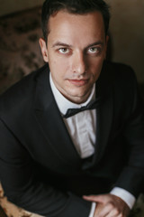 handsome Groom portrait in the morning in the room. happy man in suit with bow tie smiling, sitting in chair at window light in hotel. preparation for wedding day