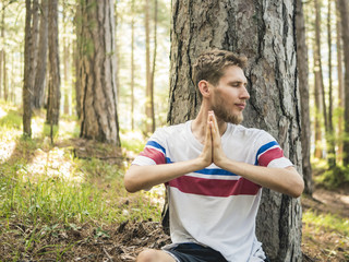 Naklejka na ściany i meble young bearded man doing yoga and meditation sitting under the tree in forest with copy space