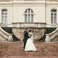 gorgeous bride with bouquet and stylish groom walking at old castle in european city street in...