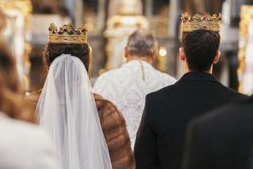 gorgeous bride and stylish groom in golden crowns, standing with priest in church during wedding...