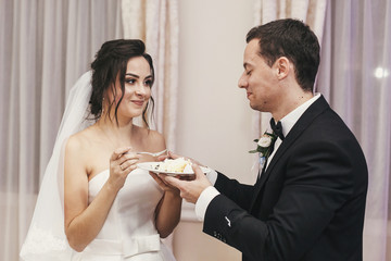gorgeous bride and stylish groom tasting their stylish wedding cake at wedding reception in restaurant. happy newlywed couple eating piece of cake, funny emotional moment.