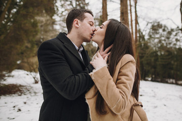 gorgeous wedding couple kissing in winter snowy park. stylish bride in coat and  groom embracing under  trees in winter forest. romantic sensual moment of newlyweds