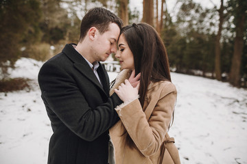 gorgeous wedding couple hugging in winter snowy park. stylish bride in coat and  groom embracing under  trees in winter forest. romantic sensual moment of newlyweds