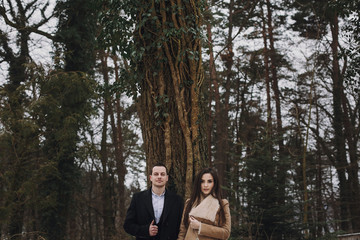 gorgeous bride in coat and stylish groom posing under trees in winter forest. happy wedding couple hugging in winter snowy park. romantic sensual moment of newlyweds