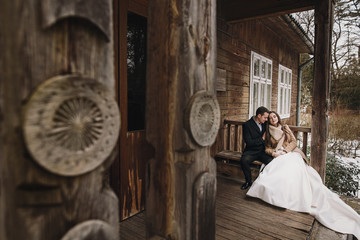 gorgeous bride in coat and stylish groom sitting at old wooden house in winter forest. happy wedding couple gently hugging on snowy porch.  barn wedding. romantic sensual moment