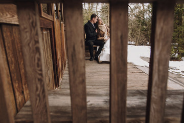 Obraz na płótnie Canvas gorgeous bride in coat and stylish groom sitting at old wooden house in winter forest. happy wedding couple gently hugging on snowy porch. barn wedding. romantic sensual moment