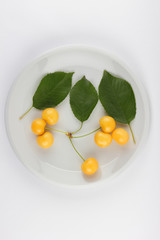 Yellow cherries with green leaves on a white plate, fresh cherries on a white background, yellow berries in minimalism style, vegetarian food, isolated
