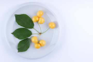 Yellow cherries with green leaves on a white plate, fresh cherries on a white background, yellow berries in minimalism style, vegetarian food, isolated