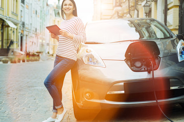 Wonderful day. Nice cheerful smiling woman standing near her amazing electric car and holding a...