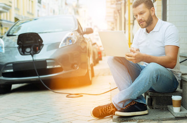 Concentrated person. Attentive serious young man sitting with a laptop not far from a charging electric car