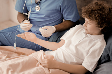 I will do it. Competent doctor sitting on the bed near ill boy and holding dropping tube in left hand while going to treat his patient