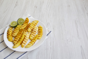 Grilled pineapple slices with lime on white plate over white wooden background, side view. Summer food. Idea for snack. Copy space and text area.
