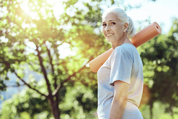 Convenient mat. Lovely smiling old woman looking calm while being in a green park with a yoga mat