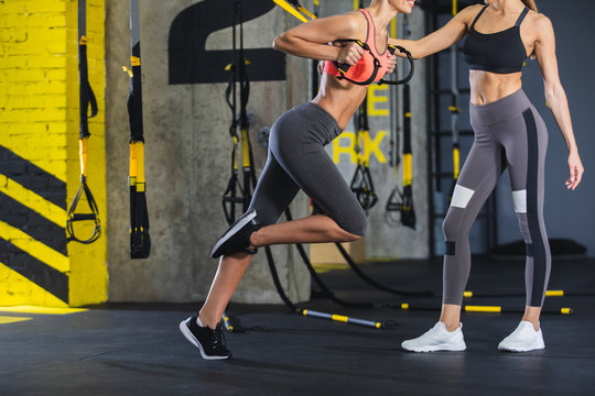 Woman coach is standing and watching sporty girl training with straps. Other slender lady is doing chest presses with trx equipment. Exercising with personal trainer in gym for better achievements
