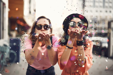 Portrait of cheerful ladies blowing confetti from hands. They situating on street during sunny day. Glad girl having fun, celebration concept