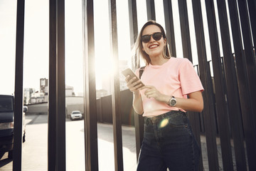 Fototapeta na wymiar Portrait of beaming girl using modern mobile while posing in urban style. She locating near iron fence on street
