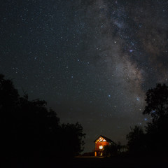 A cabin with it's windows glowing bright under the milky way at night and surrounded by the...