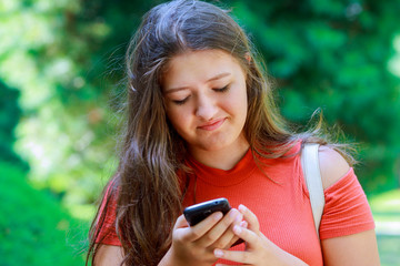 Portrait of a beautiful teen girl typing messages on the smart phone in a park with a green unfocused background