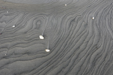 Tide patterns on the surface of a grey, sandy beach in the South Island of New Zealand. A few sea shells are scattered on the surface. 