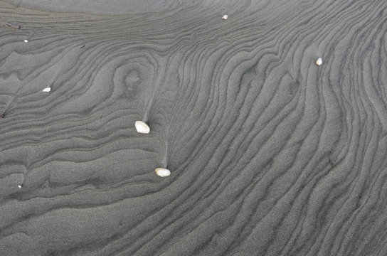 Tide patterns on the surface of a grey, sandy beach in the South Island of New Zealand. A few sea shells are scattered on the surface. 