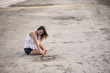 Fototapeta na wymiar one young beautiful girl, alone sitting in sand, drawing with stick, smiling. Beach, summer, casual clothing.