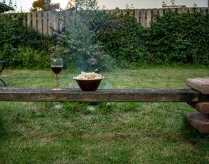 Bowl of popcorn and glass of wine stand on a bench with a campfire behind