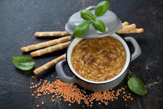 Pot of thick soup with pasta and red lentils served with grissini, horizontal shot on a brown stone background