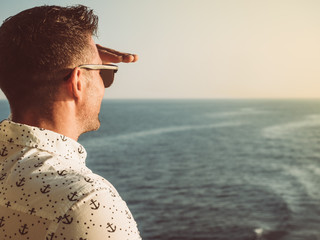 Attractive man in a white shirt with patterns in the form of anchors on the top deck of a cruise ship, looking into the distance on a sunset background. Nautical theme. Concept of sea cruises and rest