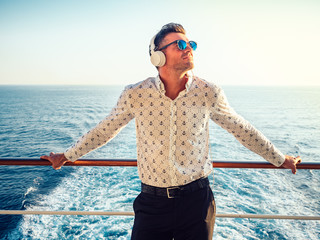 Stylish, young man on the deck of a cruise ship listening to music on white headphones against the...