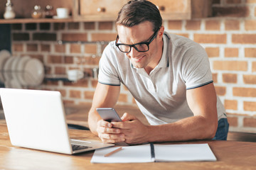 Young entrepreneur. Delighted positive nice man leaning on the table and smiling while using his...