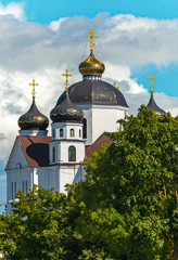 Fototapeta na wymiar A majestic church with white walls and large domes with golden crosses glistening in the sun, standing among green trees against the sky covered with dense clouds.