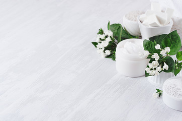 Soft pure white delicate cosmetics set of cream, salt, clay decorated white flowers, green leaves on light soft wooden background, copy space.