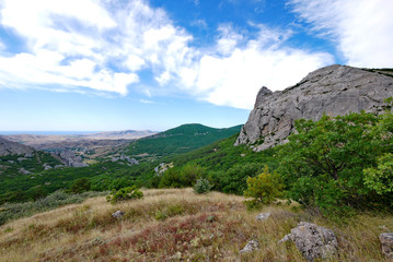 A landscape of rocky peaks and green hills against the background of azure sky with white, blurred clouds.