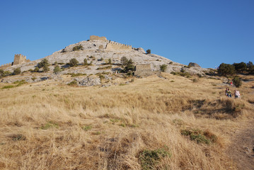 Fototapeta na wymiar Slope of a hill overgrown with dry grass with remains of crenellated walls of an ancient fortress against a blue sky and tourists descending down the path.