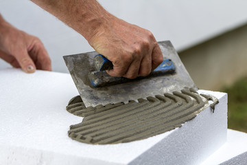 Close-up of worker hand with trowel applying glue on white rigid polyurethane foam sheet for house...
