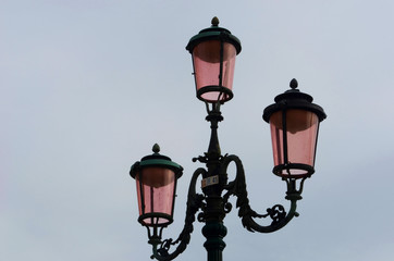 Fototapeta na wymiar A black iron lamp-post in Venice with three lamps of pink glass. They are seen against a grey sky.
