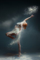 Dancing in flour concept. Redhead beauty female girl adult woman dancer in dust / fog. Girl wearing white top and shorts making dance element in flour cloud on isolated grey / black background - obrazy, fototapety, plakaty