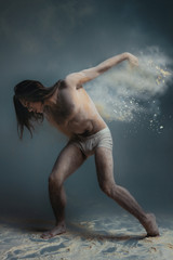 Dancing in flour concept. Long hair muscle fitness guy man male dancer in dust / fog. Guy wearing white shorts making dance element in flour cloud on isolated grey / black background
